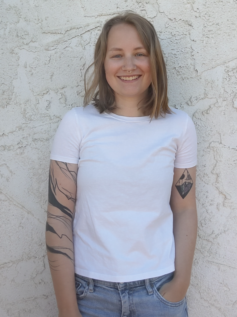 A young white woman standing against a stucco house. She has shoulder length blonde hair, a septum piercing, and tattoos.